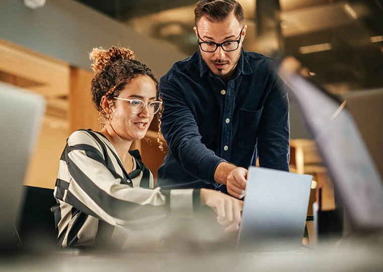 Two Colleagues Working On Laptop in Office as they Discuss a Project Two Colleagues Working On Laptop in Office as they Discuss a Project